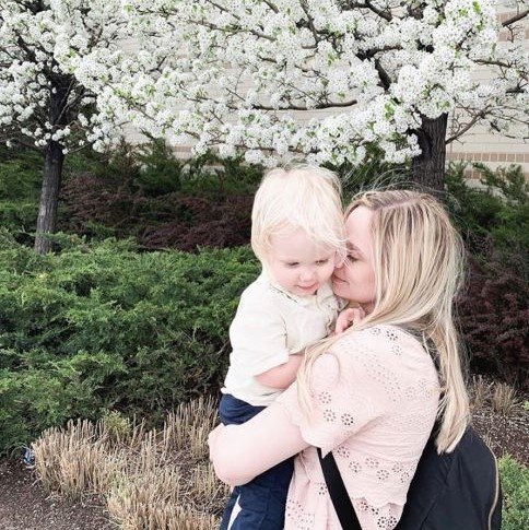 Blonde-haired woman holding toddler in front of tree with white blossoms
