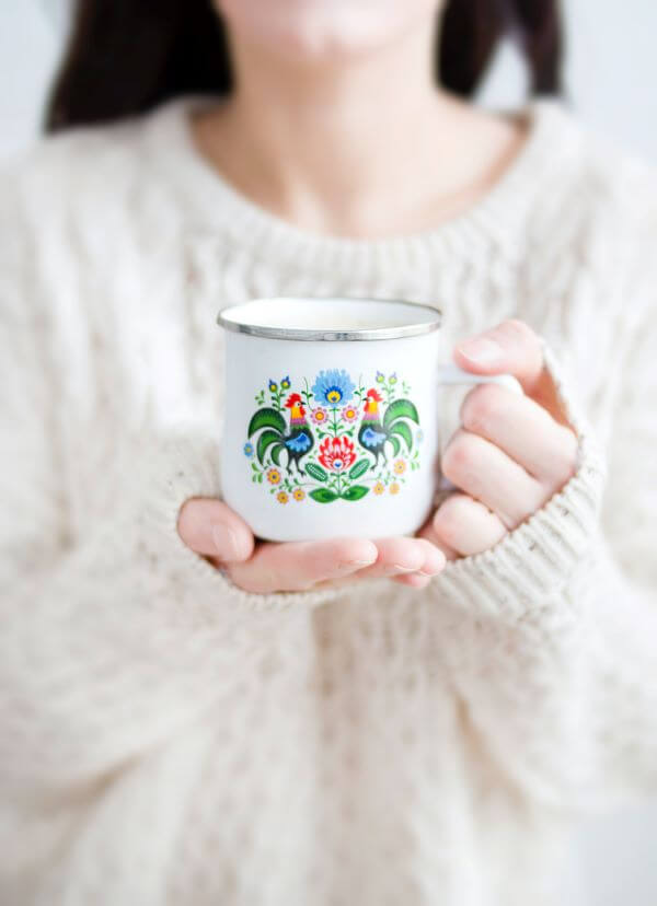 Woman in white sweater holding a white mug painted with flowers and roosters.