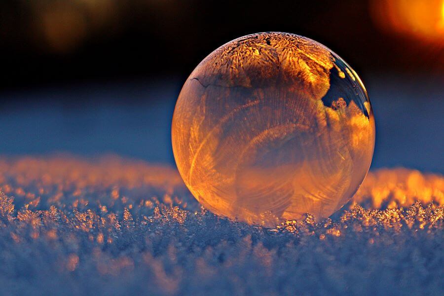 The snow on the ground appears blue-gray in the low light coming from the right side of the photo. A frozen bubble sitting on top of the snow fills most the the right side of the image. The light gives the bubble a warm, almost orange tint..