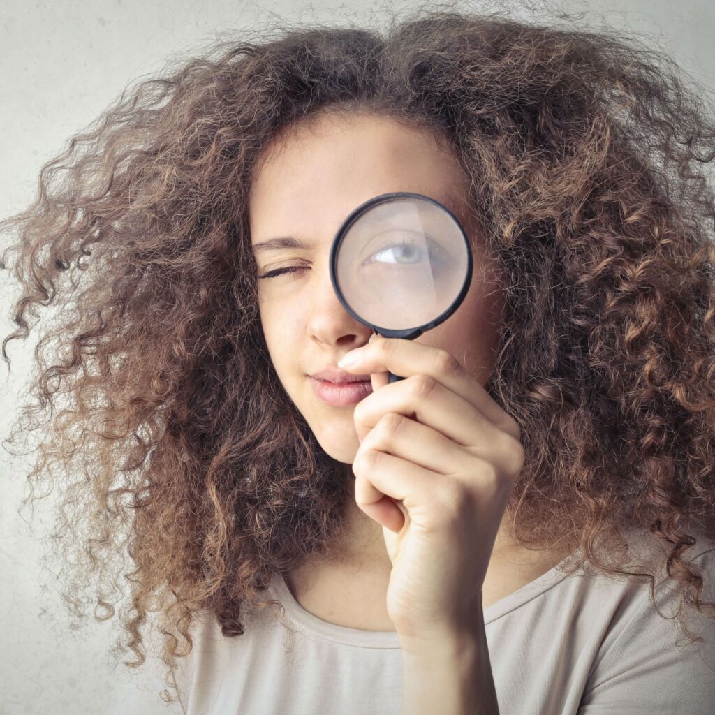 A girl with big brown curls holds a magnifying glass up to one eye. She squints to look through it towards the camera.