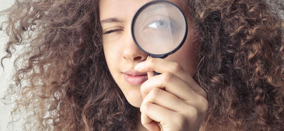 A girl with big brown curls holds a magnifying glass up to one eye. She squints to look through it towards the camera.