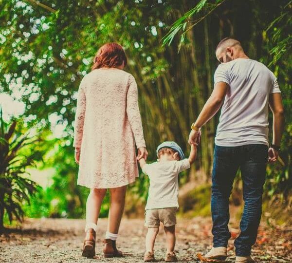 A family walks away from the camera on a path near trees. A red-haired woman wearing a dress and a man in jeans and a t-shirt hold hands with a toddler between them.