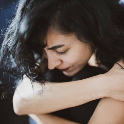 Woman with black hair sits on ground hugging her knees to her chest. She looks sad.