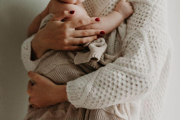 Close-up shot of a woman with red fingernail polish holding an infant in her arms. She is wearing a white sweater and the baby is wrapped in a beige blanket.