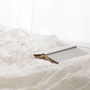 A white-covered book lies open on a rumpled white bedsheet in front of a window.