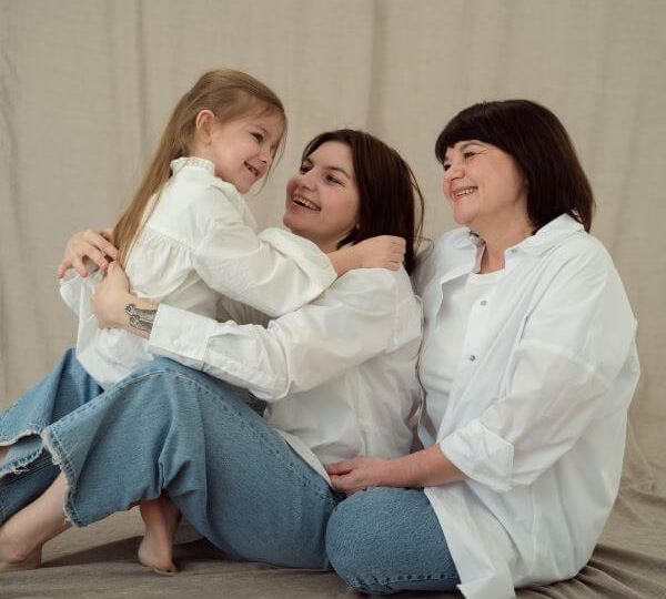 A girl with light brown hair faces her mother and grandmother, who are sitting on the floor. Each wears light wash jeans and a white button-up shirt. The mother and grandmother look lovingly at the child.